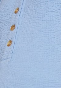 Tecido texturizado azul claro com uma pala de botões, contendo quatro botões dourados redondos. Há um detalhe de costura visível ao longo da borda.