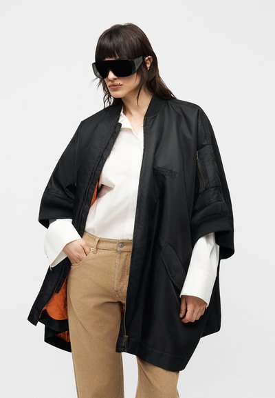 Γυναίκα που φοράει φαρδιά μαύρη μπουφάν μπόμερ πάνω από λευκό πουκάμισο και μπεζ παντελόνι, με αξεσουάρ μεγάλα μαύρα γυαλιά ηλίου, ποζάροντας μπροστά σε λευκό φόντο.