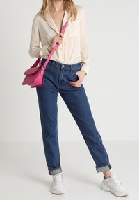 Krémová košile na knoflíky, modré džínsy s vysokým pasem, bílé tenisky a růžová crossbody taška. Hladký materiál, pohodlný střih, ohrnuté manžety.
