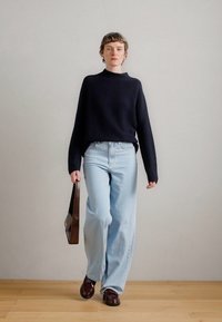 Μπλε ναυτικός πλεκτός πουλόβερ, ανοιχτό μπλε παντελόνι σε φαρδιά γραμμή και σκούρα καφέ παπούτσια. Κρατάει μια καφέ τσάντα. Μινιμαλιστικό στυλ με απλό φόντο.