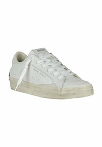 Sneaker en cuir blanc avec un bout rond, des lacets plats et une semelle légèrement texturée. Présente de subtiles perforations et une marque sur le côté.