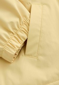 Tela de chaqueta ligera amarilla con puño de manga fruncido, que presenta una textura suave y un patrón sutil de cuadrícula. Incluye un bolsillo lateral.