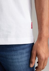 Weißes Baumwoll-T-Shirt mit abgerundetem Saum, mit einem roten Logotag auf der linken Seite, kombiniert mit blauen Jeans.