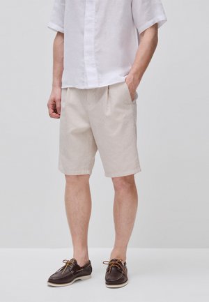 Mann trägt beige kurz gestreifte Shorts, weißes kurzärmeliges Hemd und braune Leder-Bootsschuhe, steht mit einer Hand in der Tasche.