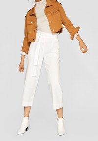 Brun, cropped denimjakke over en beige højhalset trøje, parret med hvide højhalsede bukser og hvide ankelstøvler.