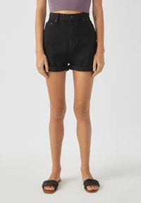 Pantalones cortos de mezclilla negra con cintura alta, puños enrollados y botones delanteros, combinados con sandalias planas negras trenzadas. Textura suave, diseño casual.