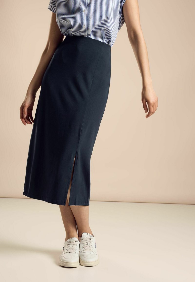 Gonna blu navy fino al ginocchio con uno spacco laterale, realizzata in tessuto morbido. Abbinata a una camicia a righe a maniche corte e scarpe da ginnastica bianche.