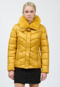Gul polstret jakke med chevron-syninger, en faux fur krave, lynlåslommer på siderne og et tætsiddende design. Båret over et sort stribet nederdel.