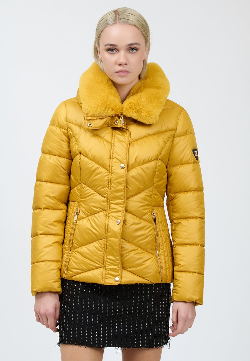 Gul polstret jakke med chevron-syninger, en faux fur krave, lynlåslommer på siderne og et tætsiddende design. Båret over et sort stribet nederdel.