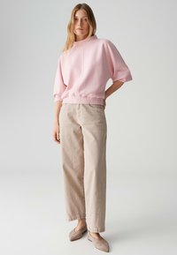 Lichtroze top met korte mouwen en een aangesloten elastische taille, gecombineerd met beige broek met wijde pijpen. Het model draagt lichtbruine instapschoenen.