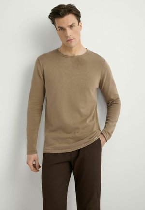 Mężczyzna ubrany w jasnobrązową koszulę z długim rękawem i ciemnobrązowe spodnie, stojący na tle gładkiej jasnej ściany, z jedną ręką w kieszeni.