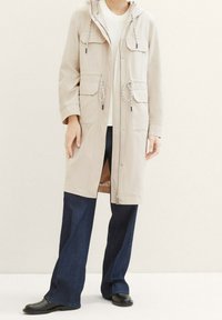 Parka impermeable beige claro con capucha, cinturilla ajustable y dos bolsillos frontales, combinado con vaqueros de pierna ancha azul oscuro y botas negras.