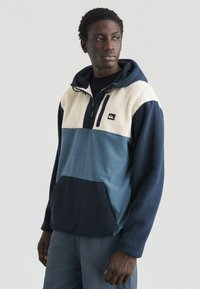 Jauns vīrietis, kurš valkā krāsu bloku flīsa kapučveida džemperi ar tumši ziliem, krējuma un ziliem paneļiem, skatoties uz sāniem pret vienkāršu baltu fonu.