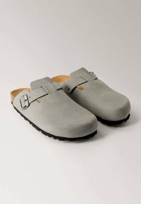 Gray suede clogs with a rounded toe, adjustable strap and buckle, and textured black rubber sole. Interior has a natural leather lining.