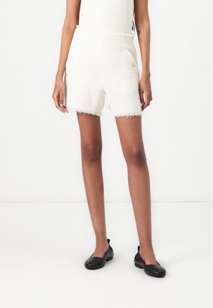 Witte textuurshorts met een rafelige zoom, twee voorzakken benadrukt door gouden knopen, gecombineerd met zwarte platte schoenen.