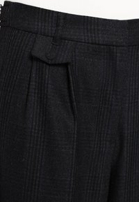 Pantalonii cu carouri negre au o linie ascuțită, un material texturat și o pliu unic la talie cu un detaliu mic, rotunjit pentru închidere.