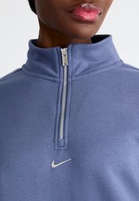 Huvtröja med dragkedja och hög krage i mjukt blått tyg. Har en silverfärgad Nike-logotyp på bröstet och metallisk detalj på dragkedjan. Slät yta hela vägen igenom.