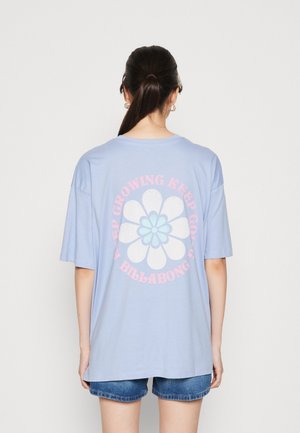 Ung kvinde med mørkt hår i en hestehale iført en lyseblå, oversized t-shirt med en blomst og teksten "Keep Growing" på ryggen samt denimshorts.