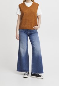 Rdzawo-pomarańczowa dzianinowa kamizelka z dekoltem w serek, zestawiona z szerokimi, wyblakłymi niebieskimi dżinsami i czarnymi sneakersami. Widoczny teksturowany wzór na kamizelce.