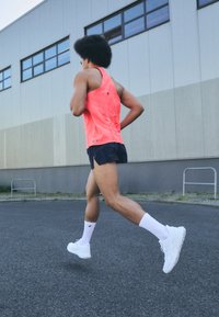 Athlet in einem leuchtend pinken, ärmellosen Oberteil und schwarzen Shorts, trägt weiße Socken und Laufschuhe, joggt auf einer gepflasterten Oberfläche.
