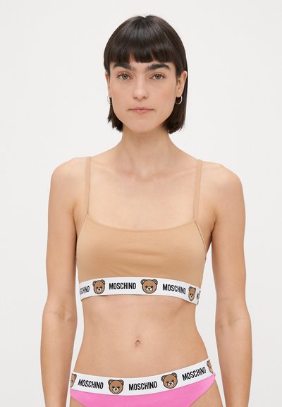 Bēšā kokvilnas krūšturis ar plānām siksni�ņām, kas ir ar baltu joslu ar lāča grafiku un tekstu "MOSCHINO". Modelei tāpat ir rozā apakšveļa.