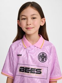 Junge Person mit langen dunklen Haaren, die ein rosa Fußballtrikot mit Kragen und der Aufschrift "BEES International" sowie dem Teamlogo auf der Vorderseite trägt.
