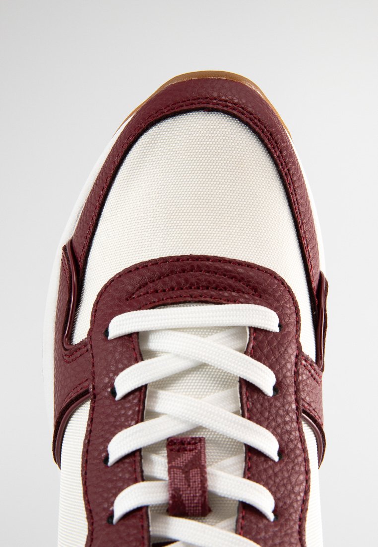 Zapatilla blanca con acentos de cuero burdeos, parte superior de lona texturizada y cordones blancos. Suela de goma visible y puntera reforzada.
