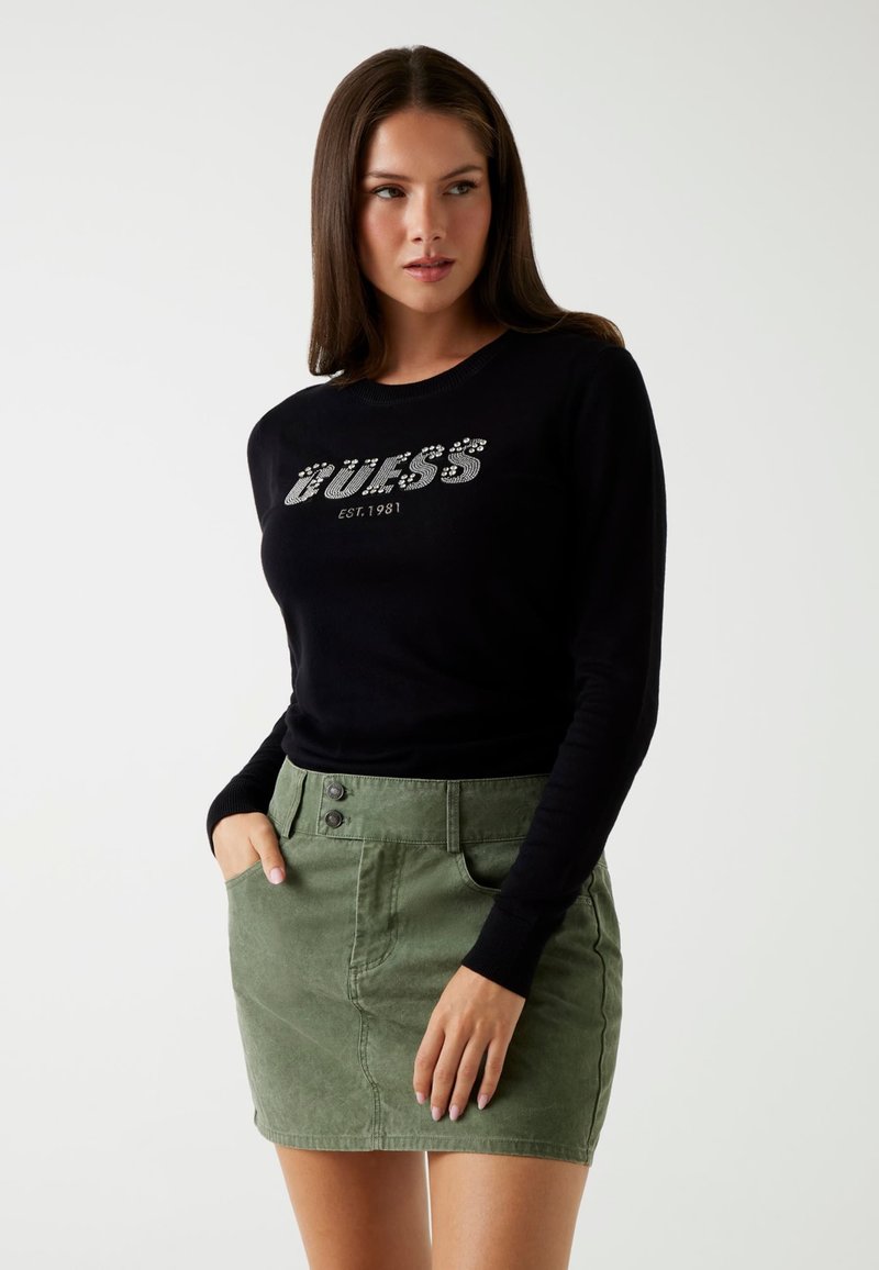 Zwarte lange mouw trui met "GUESS EST. 1981" grafiek in zilveren versiering, gecombineerd met een groene korte rok met twee knoopaccenten.