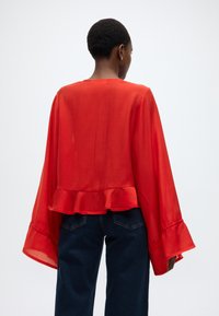 Κόκκινη, ελαφριά μπλούζα με φουσκωτό σχέδιο, που διαθέτει μακριά μανίκια και βολάν στο τελείωμα. Φορεμένη με σκούρα μπλε τζιν.