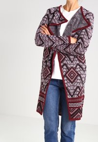 Marinröd mönstrad lång cardigan med geometrisk design, öppen front och texturerat tyg, bärs över en vit topp och blå jeans.