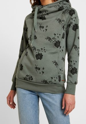 Femme portant un sweat à capuche vert olive avec des imprimés floraux et d'oiseaux noirs, associé à un jean bleu clair, debout devant un fond uni.