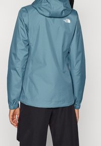 Blå vattentät jacka med huva, elastiska ärmslut och en vit logotyp på övre ryggen. Slät textur, figursydd passform och midjelång design.