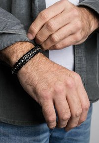 Juodas pintas odos apyrankė su sidabriniais metaliniais akcentais. Nešiojama ant riešo, dizainas pasižymi sutvirtintais tekstūromis ir saugiu uždarymu.
