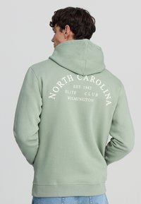 Felpa con cappuccio di colore verde chiaro, realizzata in tessuto morbido. Presenta una stampa sul retro: "NORTH CAROLINA EST. 1982 ELITE CLUB WILMINGTON" in bianco.