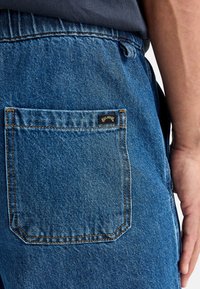 Shorts de mezclilla en azul medio con un bolsillo trasero que presenta una pequeña etiqueta de marca negra. La textura muestra la típica fabricación de jeans.