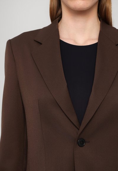 Brown blazer with a structured collar and a single button closure. Worn over a dark top, featuring smooth texture and tailored shape.