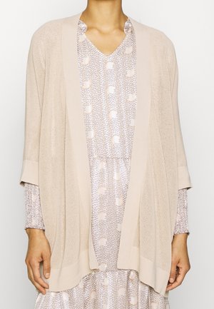 Personne portant un cardigan en maille beige ouvert sur le devant, par-dessus une robe légère à manches longues avec motif et col en V.