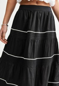 Mustat kerroksellinen hame, joka on valmistettu kevyestä kankaasta. Se sisältää röyhelöitä ja valkoista ric-rac-nauhaa saumoissa tuomaan tekstuuri- ja värieroa.