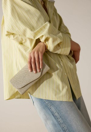 Personne portant une chemise rayée jaune clair et un jean bleu, tenant une pochette beige texturée près de la hanche.