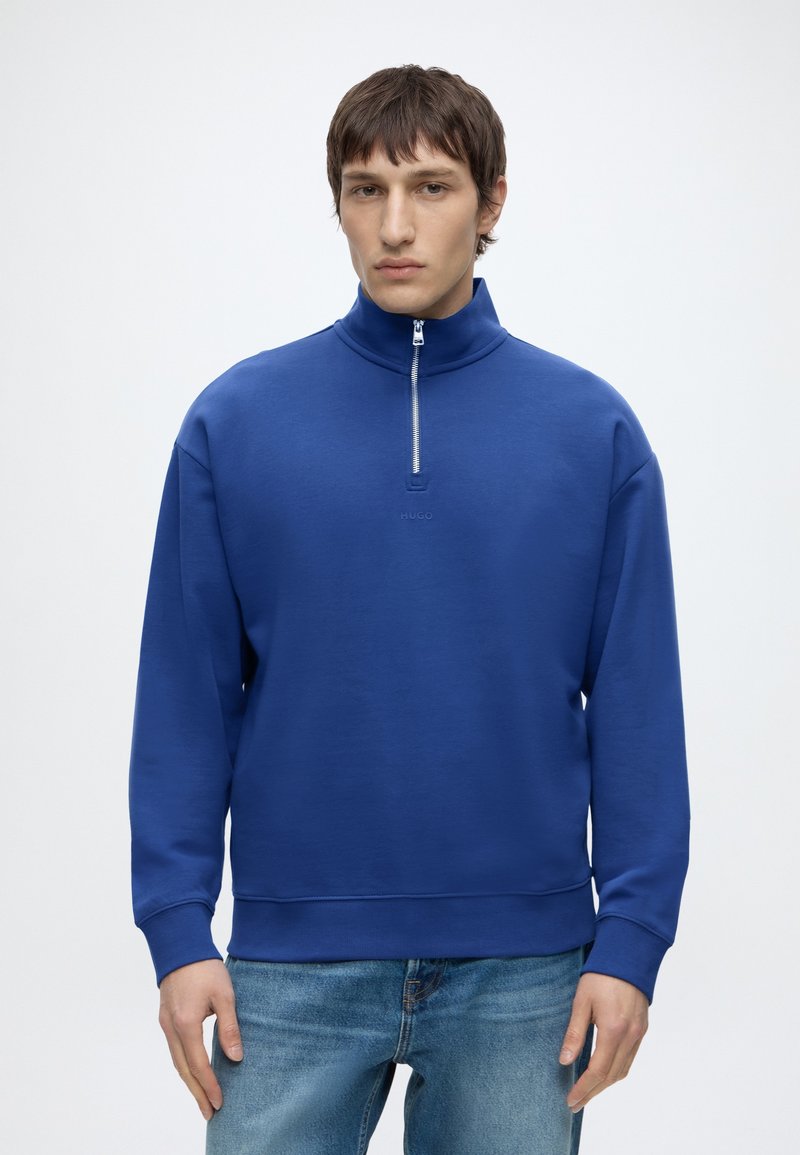 Jeune homme portant un sweat bleu demi-zip et un jean bleu clair, debout devant un fond blanc uni.