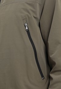 Olivgrön jacka med en vertikal svart dragkedjeficka på bröstet, något veckad tyg runt sömmen.