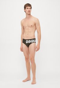Mlad moški stoji bos v črnih kopalkah blagovne znamke DSQUARED2 pred enobarvno belo ozadjem.