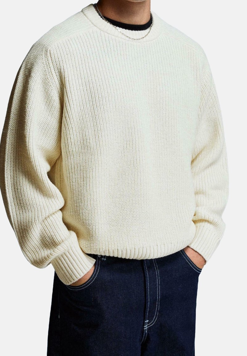 Personne portant un pull en maille côtelée ample de couleur crème et un jean bleu foncé, les mains dans les poches, sur un fond uni.
