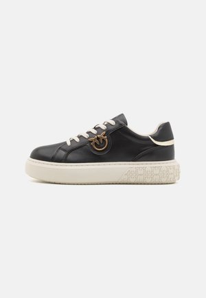 Svarte lær-sneakers med hvit såle, preget av en sirkulær gulldetalj, beige snøring og teksturert detaljering på sålen.