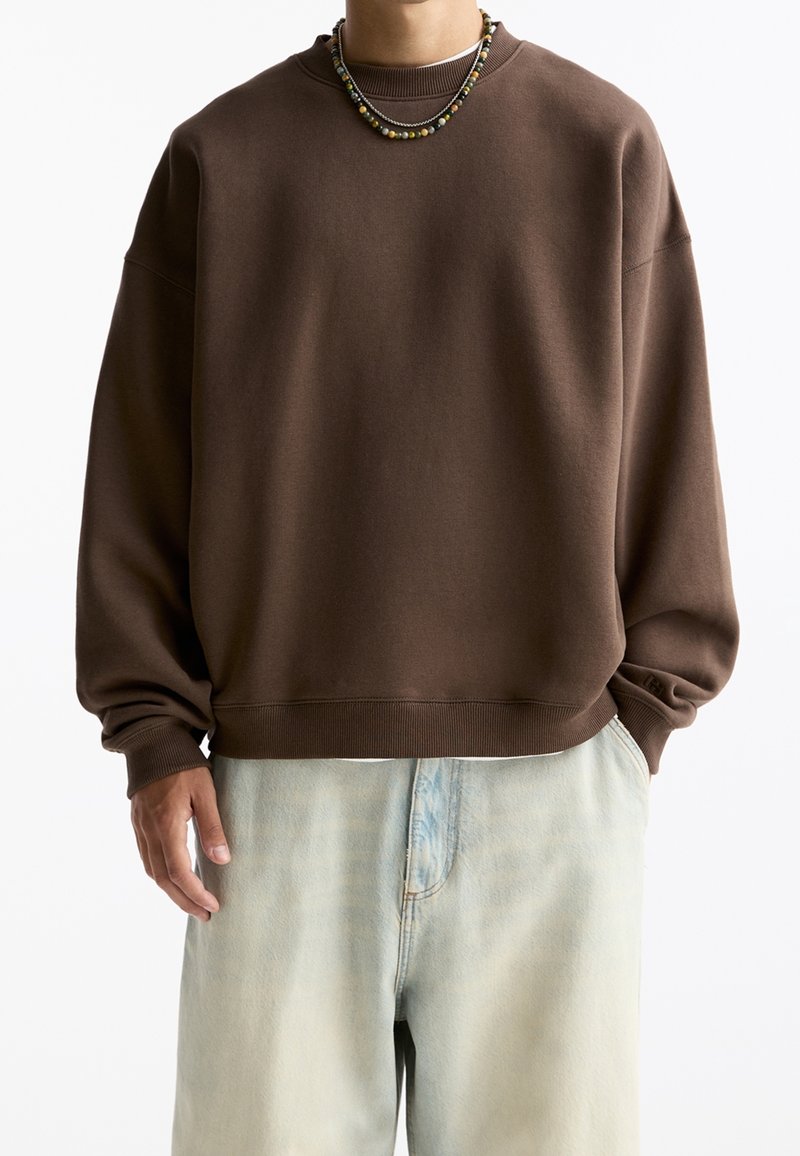 Personne portant un sweat-shirt marron oversize, un jean bleu clair délavé et deux colliers de perles, une main dans la poche et l'autre sur le côté.