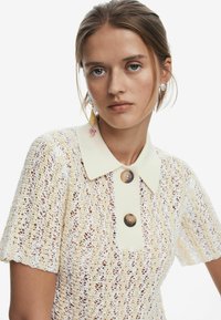 Pletený top v krémovej a bielej farbe s golierom, ktorý má veľké hnedé gombíky a krátke rukávy, ukazujúci textúrovaný čipkovaný vzor.