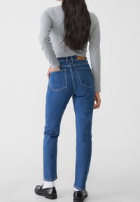 Jeans in denim blu a vita alta con gamba dritta, dotati di due tasche posteriori e di un'etichetta in pelle sulla vita. Indossati con una camicia grigia a maniche lunghe.