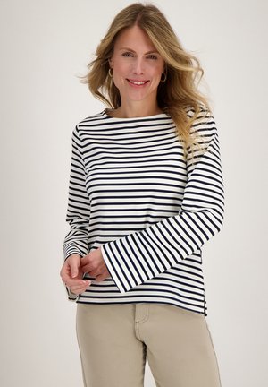 Vrouw met lang blond haar die een wit en marineblauw gestreepte top met lange mouwen en beige broek draagt, glimlachend met gevouwen handen.