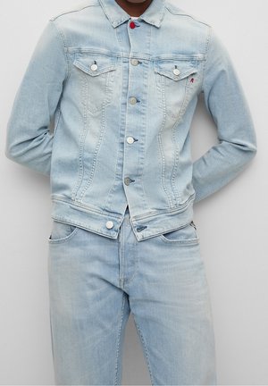 Muž v světle modré džínové bundě zapnuté do knoflíků, ve stejných světle modrých džínách, s rukama za zády, stojící před jednobarevným pozadím.