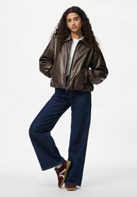 Veste bomber en cuir synthétique marron avec fermeture éclair, top blanc, jeans évasés bleus et baskets bourgogne avec accents blancs. Tenue décontractée.